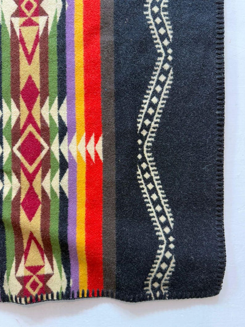 May include: A woven blanket featuring a geometric pattern with vertical stripes in red, yellow, purple, green, and cream. The right side displays a large black section with a cream-coloured wavy line pattern and a finished stitched edge.