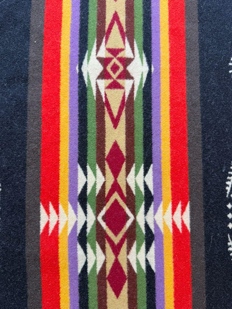 May include: A textile featuring a vertical pattern with a central column of geometric diamond and arrow shapes in maroon, tan, and white. This is flanked by symmetrical stripes of green, black, purple, yellow, and red on a dark navy blue background.