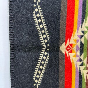 May include: A wool blanket featuring a black base with a cream-coloured geometric wave pattern along the edge. The design includes vertical stripes in red, brown, yellow, purple, and green, accented by a repeating triangular motif in cream and green.
