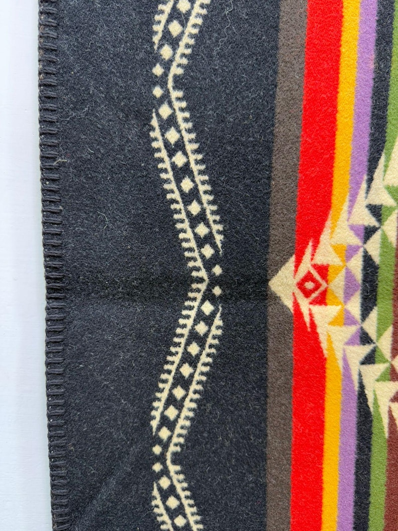 May include: A wool blanket featuring a black base with a cream-coloured geometric wave pattern along the edge. The design includes vertical stripes in red, brown, yellow, purple, and green, accented by a repeating triangular motif in cream and green.