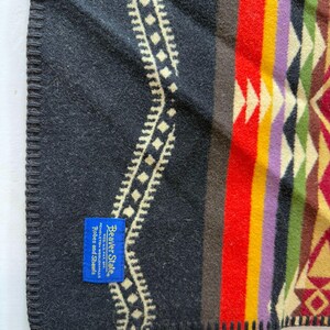 May include: A black blanket with a geometric pattern in red, yellow, purple, and green. A blue rectangular tag is sewn onto the corner with the text Beaver State, REG. U.S. PAT. OFF., PENDLETON WOOLEN MILLS, Robes and Shawls. The edge is stitched.