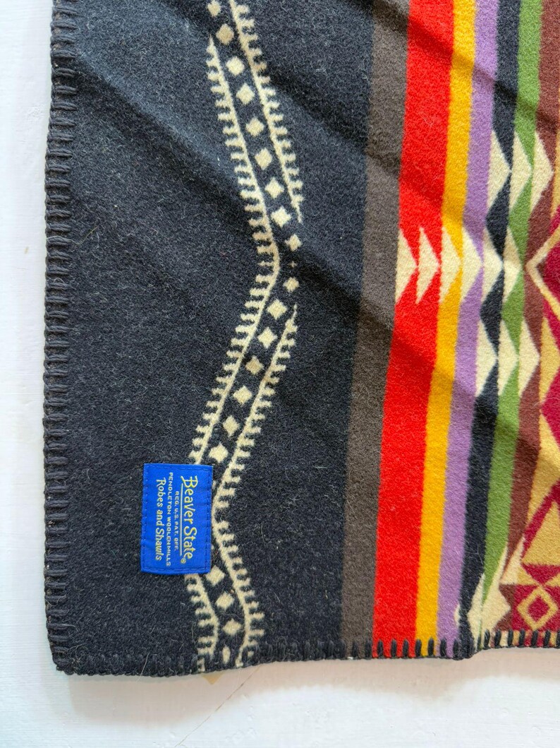 May include: A black blanket with a geometric pattern in red, yellow, purple, and green. A blue rectangular tag is sewn onto the corner with the text Beaver State, REG. U.S. PAT. OFF., PENDLETON WOOLEN MILLS, Robes and Shawls. The edge is stitched.