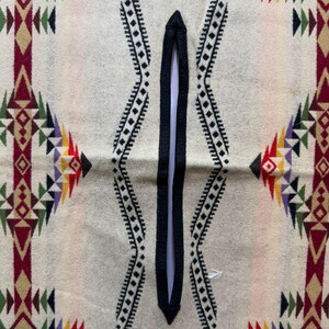 May include: A cream-coloured poncho featuring a central black vertical neck opening. The fabric displays a repeating geometric pattern in shades of maroon, dark green, yellow, and purple, arranged in vertical bands and symmetrical diamond shapes.