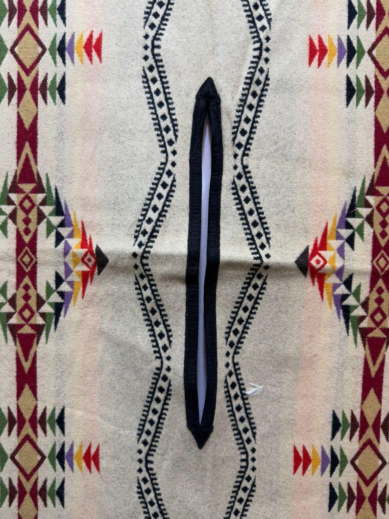 May include: A cream-coloured poncho featuring a central black vertical neck opening. The fabric displays a repeating geometric pattern in shades of maroon, dark green, yellow, and purple, arranged in vertical bands and symmetrical diamond shapes.