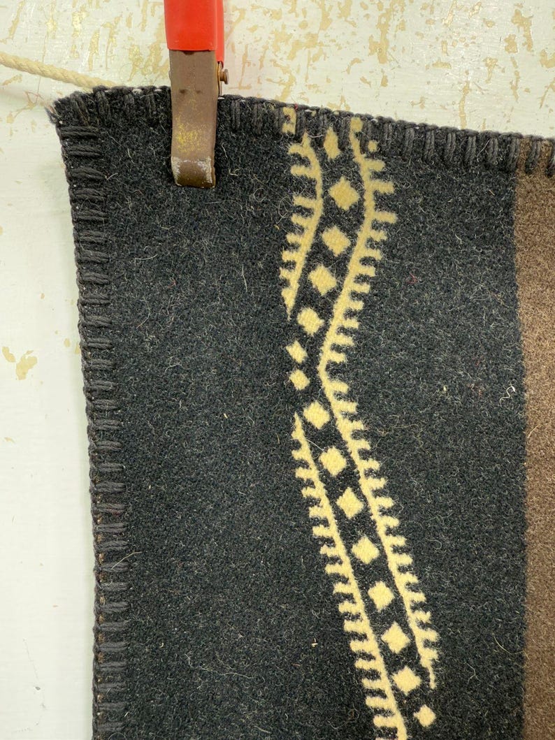 May include: A thick, dark charcoal grey wool blanket featuring a vertical, wavy cream-coloured geometric pattern of diamonds and serrated edges. The edge of the fabric is finished with a dark, heavy blanket stitch, and a brown section is visible.