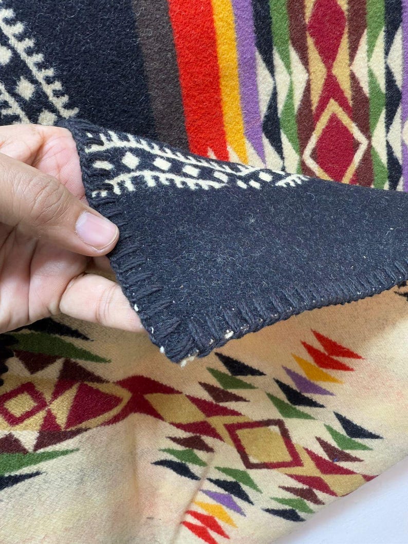 May include: A thick, soft textile blanket featuring a geometric pattern in black, cream, red, yellow, green, and purple. The fabric has a dark black underside with a visible whip-stitched edge, contrasting with the lighter cream-coloured top side.