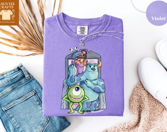May include: A violet Comfort Colors sweatshirt featuring a graphic of characters from the movie Monsters, Inc. The design includes Mike Wazowski, Sully, and Boo. A silver necklace with a charm is on the shirt. The shirt is next to blue jeans and white shoes.