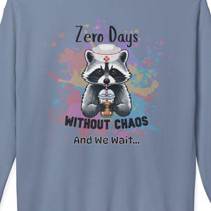May include: A light blue sweatshirt featuring a raccoon wearing a nurse's hat and holding a beverage. The text on the sweatshirt reads "Zero Days Without Chaos" and "And We Wait..." in a playful font, with colorful paint splatters.