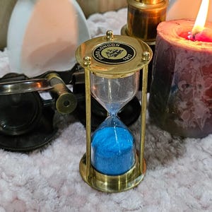 Puede incluir: Un reloj de arena de latón con arena azul, marcado "ROSS LONDON 1917". El reloj de arena se encuentra frente a una vela encendida, un pequeño telescopio y objetos decorativos, creando una estética vintage.