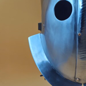 May include: A close-up of a silver metal helmet with a circular eye opening. The helmet has a curved visor and is mounted on a black stand. The surface of the helmet shows signs of wear.