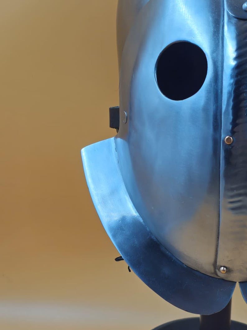 May include: A close-up of a silver metal helmet with a circular eye opening. The helmet has a curved visor and is mounted on a black stand. The surface of the helmet shows signs of wear.