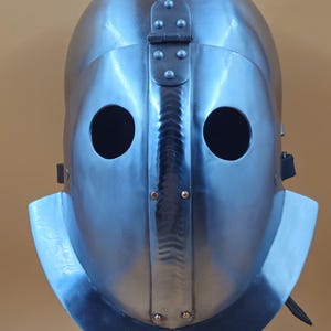 May include: A steel helmet with a polished finish, featuring two circular eye openings and a hinged top section. The helmet's design includes a central ridge and a flared lower edge. The metal surface reflects light, showing highlights and shadows.