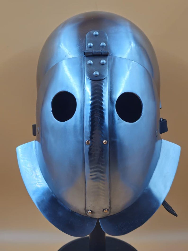 May include: A steel helmet with a polished finish, featuring two circular eye openings and a hinged top section. The helmet's design includes a central ridge and a flared lower edge. The metal surface reflects light, showing highlights and shadows.