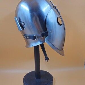 May include: A steel helmet with a rounded crown and a hinged face guard, displayed on a black stand. The helmet has a circular eye opening and is likely for historical reenactment or theatrical purposes. The metal has a polished finish.