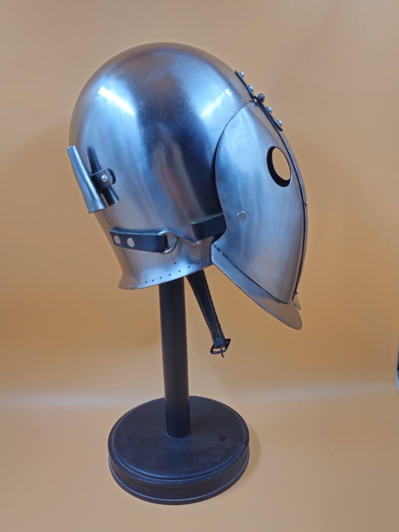 May include: A steel helmet with a rounded crown and a hinged face guard, displayed on a black stand. The helmet has a circular eye opening and is likely for historical reenactment or theatrical purposes. The metal has a polished finish.