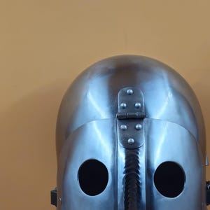 May include: A shiny, silver-coloured medieval helmet with two eye holes and a hinged visor. The helmet is made of metal and has a smooth, reflective surface. The background is a solid, muted orange.