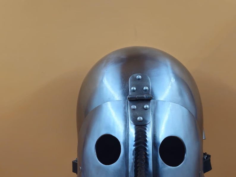 May include: A shiny, silver-coloured medieval helmet with two eye holes and a hinged visor. The helmet is made of metal and has a smooth, reflective surface. The background is a solid, muted orange.