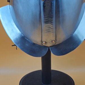 May include: A close-up of a polished steel helmet with a flared neck guard. The helmet is mounted on a black stand against a light brown background. The helmet has rivets and a central ridge.