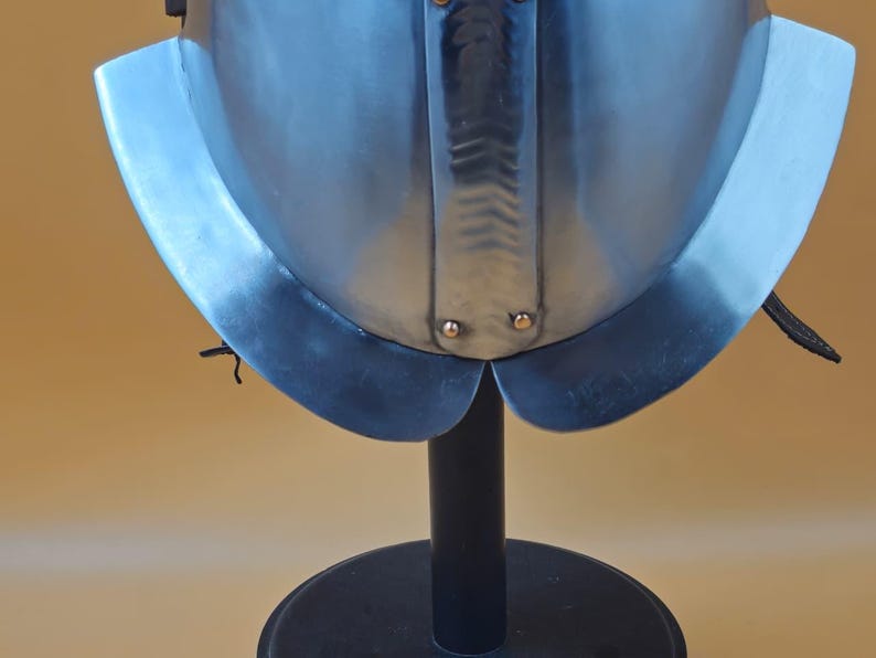 May include: A close-up of a polished steel helmet with a flared neck guard. The helmet is mounted on a black stand against a light brown background. The helmet has rivets and a central ridge.