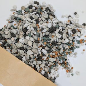 May include: A collection of mixed-colour horticultural pebbles, including white, grey, black, green, and orange stones. The pebbles vary in size and texture, suitable for plant potting mixes. A brown paper bag is partially visible in the lower left corner.