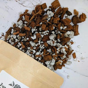 Peut inclure: Gros plan d'un mélange de copeaux de coque de noix de coco bruns et de perlite blanche. Le mélange est dispersé sur une surface blanche, avec un sac en papier brun partiellement visible dans le coin inférieur gauche. Le mélange est pour les plantes.