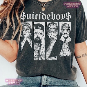 Puede incluir: Camiseta gris oscuro con el nombre de la banda "Suicideboys" en fuente gótica. Debajo, un gráfico de cuatro miembros de la banda en paneles separados, con tatuajes y sombreros. La camiseta tiene un aspecto vintage y desgastado.