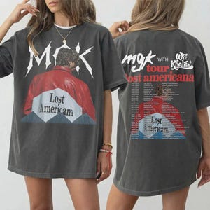 Può includere: T-shirt grigio scuro con grafica di una persona con giacca rossa e la scritta "Lost Americana". La parte anteriore della maglietta presenta le lettere "MGK" in bianco. Il retro della maglietta presenta le date del tour e la scritta "mgk with Wiz Khalifa tour Lost Americana."