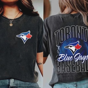 Puede incluir: Camiseta gris oscuro con el logotipo de los Blue Jays de Toronto en la parte delantera y trasera. La parte delantera muestra el logotipo del equipo, un arrendajo azul con una hoja de arce roja. La parte trasera tiene las palabras "TORONTO Blue Jays BASEBALL" sobre una pelota de béisbol.