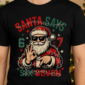 Puede incluir: Camiseta negra con un diseño de Papá Noel con gafas de sol y traje rojo, rodeado de luces navideñas. El texto "SANTA SAYS" y "SIX SEVEN" está en rojo y verde.