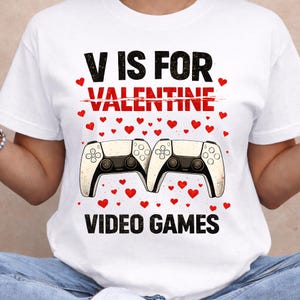 May include: White t-shirt with the text "V IS FOR VALENTINE" with a line through "VALENTINE" and the text "VIDEO GAMES". The shirt features two game controllers and red heart graphics.