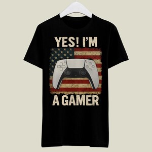 Könnte beinhalten: Schwarzes T-Shirt mit dem Text "YES! I'M A GAMER" über einem weißen und schwarzen Gamecontroller-Design. Der Controller befindet sich vor einem verwitterten Hintergrund der amerikanischen Flagge.