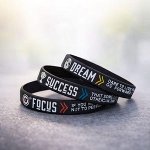 May include: Three black rubber wristbands with white, yellow, and red text. The bands read "FOCUS", "SUCCESS", and "DREAM". Each band has a different motivational message and graphic.