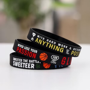 May include: Three black silicone wristbands stacked on top of each other. Each band has a different motivational message in white, red, yellow, and green. One band has a basketball graphic.