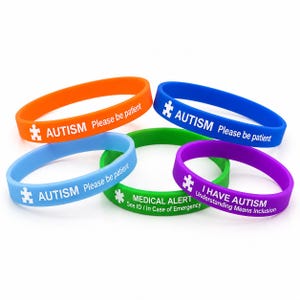 Op de afbeelding: Vijf siliconen polsbandjes in oranje, blauw, lichtblauw, groen en paars. De oranje, blauwe en lichtblauwe bandjes hebben een puzzelstuk-icoon en de tekst "AUTISM Please be patient". Het groene bandje zegt "MEDICAL ALERT See ID / In Case of Emergency". Het paarse bandje zegt "I HAVE AUTISM Understanding Means Inclusion".