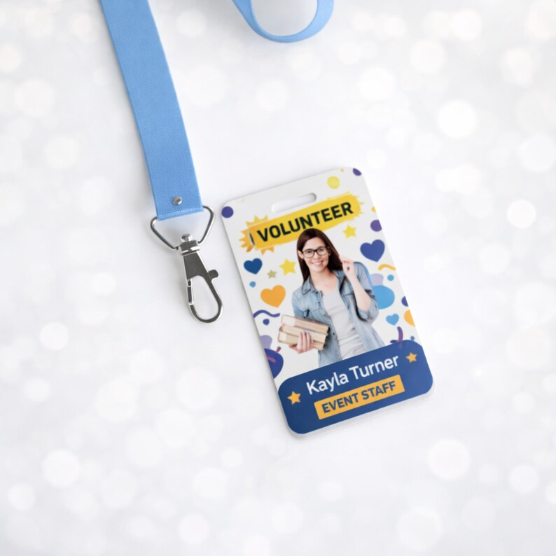 May include: A light blue lanyard with a silver metal clip attached to a rectangular ID badge. The badge features a white background with yellow, blue, and purple shapes, the text VOLUNTEER, a photo of a person holding books, and Kayla Turner EVENT STAFF.