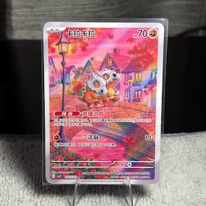 May include: A collectible trading card featuring two orange Cubone characters with bone helmets sitting on a brick bridge in a town at sunset. The card displays the name "卡拉卡拉", 70 HP, a fighting energy symbol, and Chinese text detailing abilities and attacks.