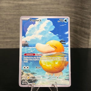 Puede incluir: Una carta coleccionable de Pokémon Psyduck. La tarjeta presenta una imagen de dibujos animados de Psyduck en el agua. La tarjeta tiene un fondo azul y blanco con el texto "Psyduck" y "70 HP". La tarjeta se muestra en un soporte de plástico transparente.