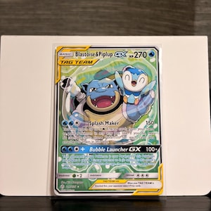 Peut inclure: Une carte à collectionner Pokémon avec Blastoise et Piplup. La carte présente un schéma de couleurs bleu et blanc avec le texte "Blastoise & Piplup GX" et "TAG TEAM". La carte comprend également le texte "Splash Maker" et "Bubble Launcher GX".