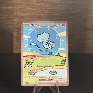 Op de afbeelding: Een Pokémon Mew ex ruilkaart. De kaart toont een lichtblauw Mew-personage in een blauwe cirkel met sterren. De kaart heeft de tekst "Restart" en "Genome Hacking". De kaart wordt weergegeven op een transparante plastic standaard.