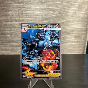 May include: A Mega Charizard X EX Pokémon trading card. The card features a black and blue Charizard illustration with the text "Inferno X". The card is displayed on a clear plastic stand.