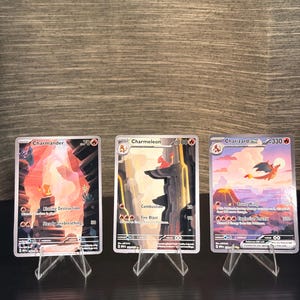 May include: Three Pokemon trading cards, featuring Charmander, Charmeleon, and Charizard EX, are displayed on clear acrylic stands. The cards have colorful illustrations of the characters in a landscape setting. The Charizard card shows a volcano and a flying dragon.