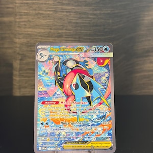 May include: A holographic Mega Greninja EX Pokémon trading card. The card features a blue and black creature with red accents, and the text "Mega Greninja EX" and "Ninja Spinner". The card is displayed on a clear plastic stand.