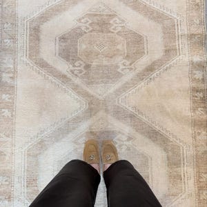 Op de afbeelding: Een rechthoekig vloerkleed in neutrale beige en lichtbruine tinten met een centraal geometrisch ruitpatroon en een decoratieve rand. Het kleed heeft een laagpolige textuur en toont symmetrische, tribaal geïnspireerde motieven.