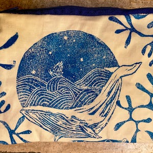 Può includere: Una pochette rettangolare con cerniera in tessuto color avorio con un disegno serigrafato blu. La grafica raffigura una grande balena che nuota tra le onde sotto un cielo stellato con una piccola nave, circondata da motivi simili a coralli.