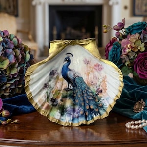 May include: A large, white seashell with a gold-painted rim and a hand-painted peacock design. The peacock is depicted in shades of blue and green, surrounded by floral accents. A decorative item for home decor.