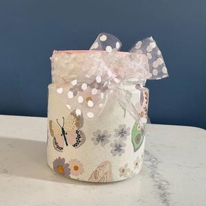 May include: A cylindrical jar decorated with butterflies and flowers in pastel colours. The jar is wrapped with a white and pink fabric and topped with a sheer, polka-dotted bow. The jar sits on a white surface against a dark blue background.