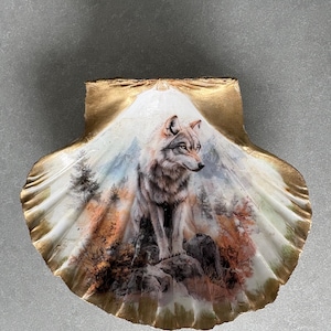 May include: A large scallop shell featuring a detailed painting of a grey wolf standing on rocks in a forest landscape. The shell has a scalloped edge painted with a metallic gold finish, framing the central wildlife artwork in natural tones.