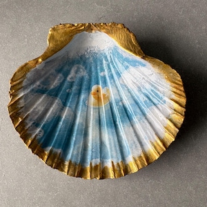 May include: A decorative scallop shell featuring a hand-painted scene of a yellow rubber duck floating in blue water with white clouds. The edges of the shell are painted with a metallic gold finish, highlighting the natural ridges and texture.