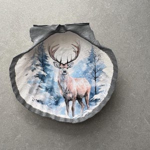 May include: A decorative seashell with a painted winter scene. The artwork features a majestic deer standing in a snowy forest with blue and white trees. The shell has a grey border.