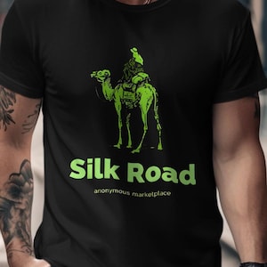 May include: Black t-shirt with a green illustration of a camel and rider. The text "Silk Road" is printed in green, with "anonymous marketplace" underneath. The shirt is made of a soft material.